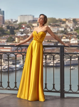 Robe de cocktail longue en satin jaune d'or avec bustier cœur drapé et ceinture fine, portée dans un cadre urbain avec vue sur un port.