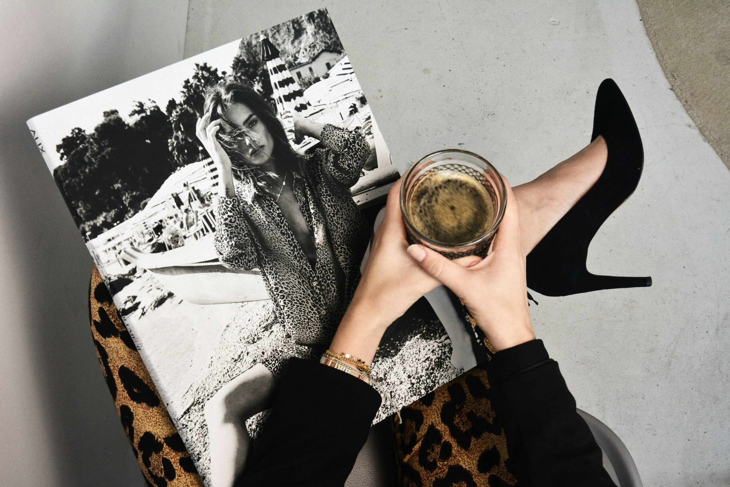 Femme tenant un verre de champagne, avec un magazine en noir et blanc posé sur ses genoux, portant des chaussures à talons noirs et un pantalon imprimé léopard.