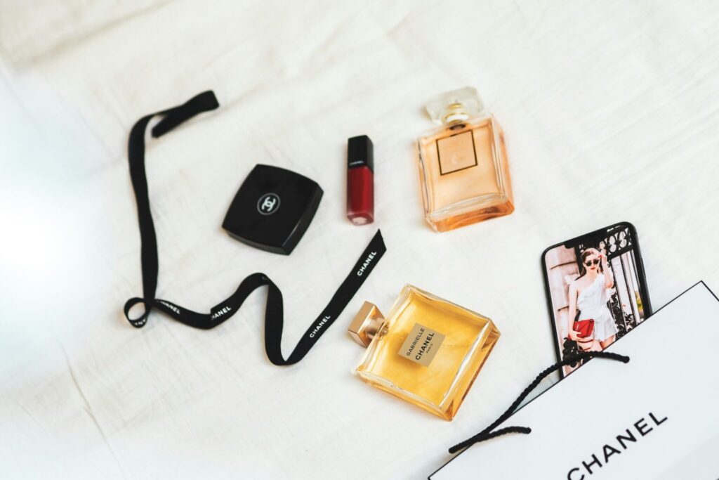 Accessoires de mode comprenant des parfums, du rouge à lèvres, une poudre compacte et un collier noir, disposés sur un fond blanc avec un sac Chanel.