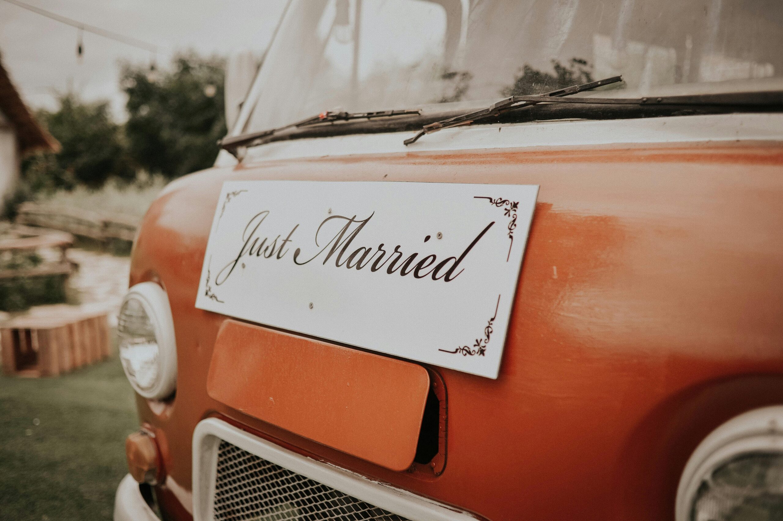 Avant d’un van vintage orange avec une pancarte 'Just Married' accrochée, dans un décor extérieur champêtre.