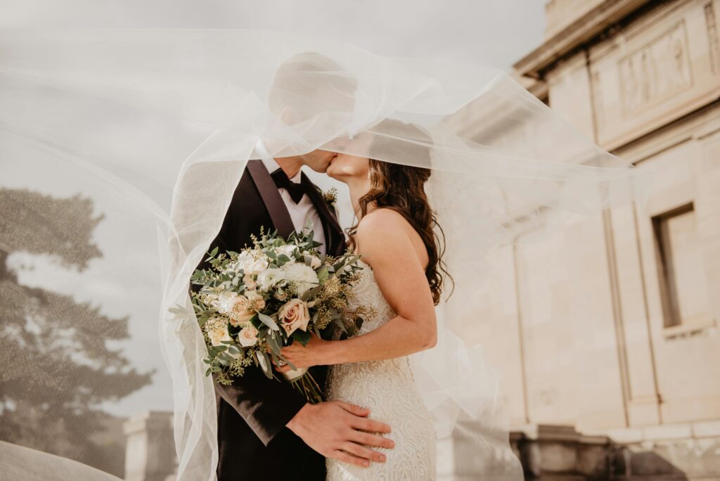 Couple de mariés s'embrassant sous un voile transparent, la mariée tenant un bouquet de fleurs pastel, avec un bâtiment historique en arrière-plan.
