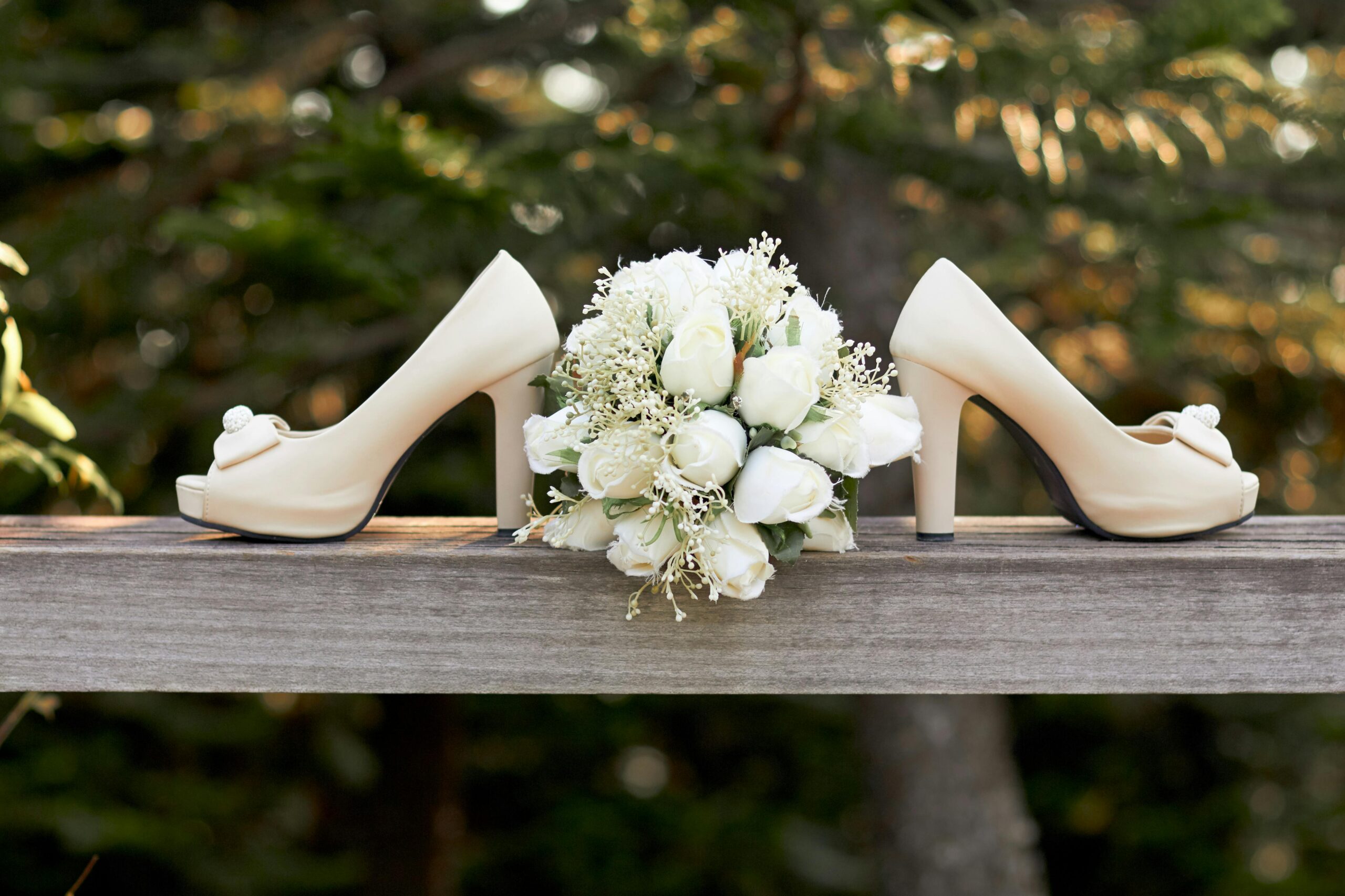 Paire de talons beige posée sur une barrière en bois avec un bouquet de fleurs blanches au centre, dans un cadre naturel.