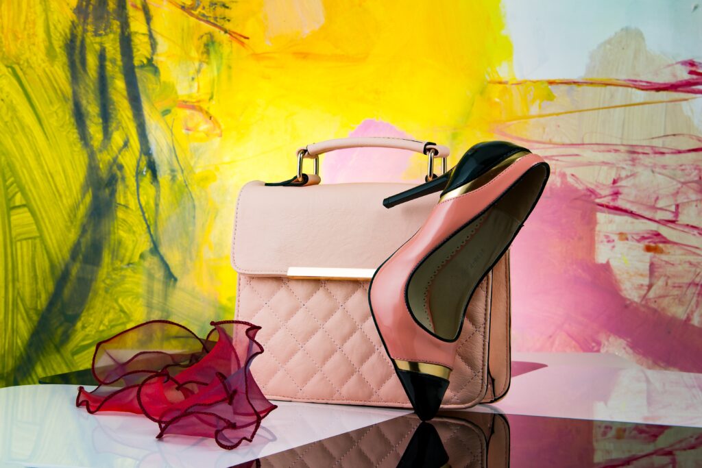 Sac à main beige matelassé accompagné d'une chaussure à talon rose et noir, posé sur une surface réfléchissante avec un fond artistique jaune et rose