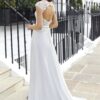 Vue de dos d'une robe de mariée blanche avec un décolleté plongeant et une rangée de boutons perlés, portée par une femme sur des marches devant une clôture en fer forgé