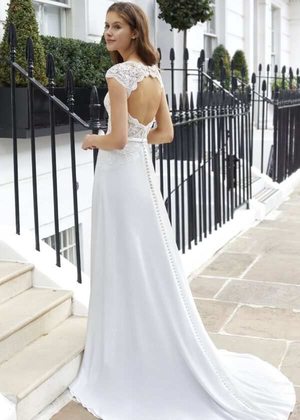 Vue de dos d'une robe de mariée blanche avec un décolleté plongeant et une rangée de boutons perlés, portée par une femme sur des marches devant une clôture en fer forgé