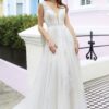Une mariée souriante posant dans une élégante robe blanche avec des détails de dentelle, un décolleté plongeant et une jupe fluide. La scène est capturée devant des maisons pastel, avec une touche de charme urbain
