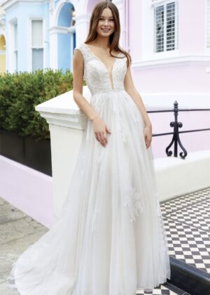 Une mariée souriante posant dans une élégante robe blanche avec des détails de dentelle, un décolleté plongeant et une jupe fluide. La scène est capturée devant des maisons pastel, avec une touche de charme urbain
