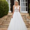 Une mariée souriante portant une robe blanche avec un corsage en dentelle florale, un décolleté en V et une jupe fluide en tulle, posant devant une villa élégante.