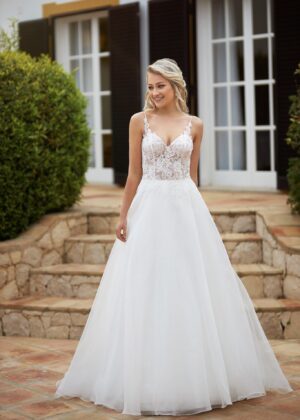 Une mariée souriante portant une robe blanche avec un corsage en dentelle florale, un décolleté en V et une jupe fluide en tulle, posant devant une villa élégante.
