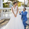 Une mariée de dos en robe blanche avec un dos nu orné de dentelle florale et une longue traîne fluide, posant sur des marches en pierre dans un jardin décoré de statues.