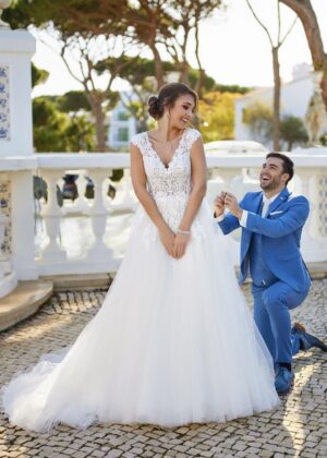 Une mariée de dos en robe blanche avec un dos nu orné de dentelle florale et une longue traîne fluide, posant sur des marches en pierre dans un jardin décoré de statues.