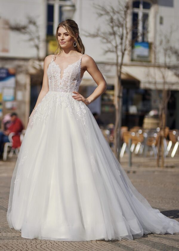 Mariée en robe blanche à décolleté en V profond, corsage en dentelle, jupe volumineuse fluide ornée de détails scintillants, posant dans une place urbaine sous un ciel ensoleillé
