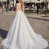 Vue arrière d’une mariée portant une robe de mariée à dos nu en dentelle, avec une jupe volumineuse fluide et une longue traîne, dans une ambiance urbaine avec des passants et cafés en arrière-plan