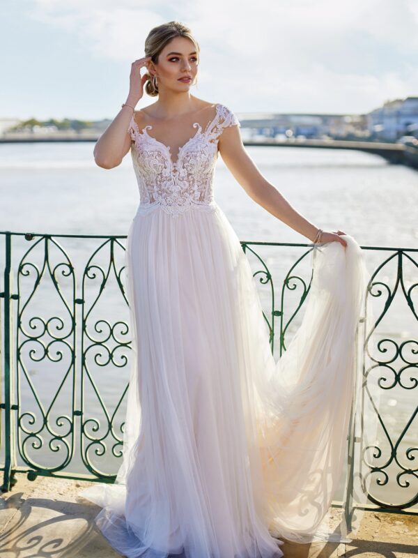 Mariée élégante en robe de mariée fluide avec un corsage en dentelle délicate, posant sur un balcon au bord de l’eau, sous un ciel clair