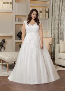 Mariée debout dans une robe de mariée blanche avec des détails en dentelle et une jupe volumineuse. Elle se tient dans un salon élégant, entourée de meubles raffinés et d'une décoration chaleureuse