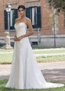 Mariée élégante dans une robe bustier scintillante ornée de motifs floraux, posant dans un jardin verdoyant avec une architecture classique en arrière-plan