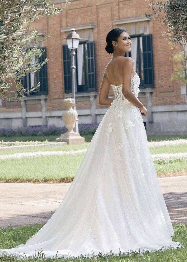 Une mariée porte une robe de mariée au style princesse avec un bustier sans bretelles orné de broderies florales délicates et une longue traîne en tulle scintillant. Elle est debout dans un jardin verdoyant avec un bâtiment en briques rouges en arrière-plan, offrant une ambiance romantique et élégante