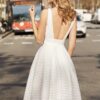 Vue arrière d'une mariée portant une robe mi-longue élégante en tissu texturé blanc, mettant en valeur un décolleté plongeant dans le dos. La robe est cintrée à la taille, créant une silhouette flatteuse. La scène se déroule dans une rue urbaine, avec des éléments de transport en arrière-plan, soulignant une ambiance citadine moderne