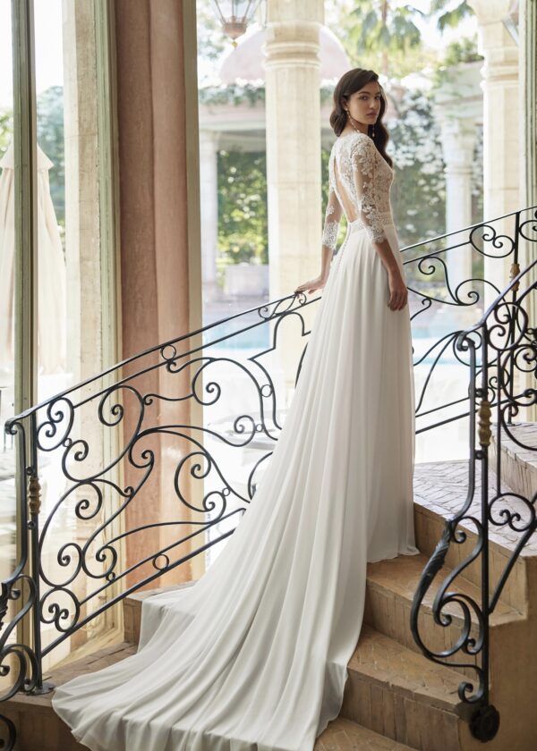 Vue arrière d'une mariée élégante portant une robe en dentelle avec dos ouvert et une longue traîne fluide, sur un escalier orné de fer forgé