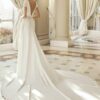 Mariée de dos en robe de mariée avec décolleté plongeant et longue traîne, debout dans une salle avec des colonnes et de grandes fenêtres