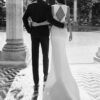 Vue arrière d’un couple marchant bras dessus bras dessous, la mariée portant une robe avec un dos ouvert triangulaire et le marié en costume élégant