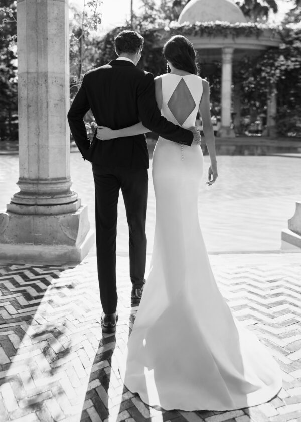 Vue arrière d’un couple marchant bras dessus bras dessous, la mariée portant une robe avec un dos ouvert triangulaire et le marié en costume élégant