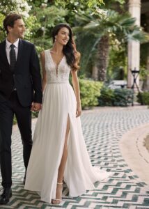 Couple souriant marchant main dans la main, la mariée portant une robe fluide avec un décolleté en V en dentelle et une fente élégante