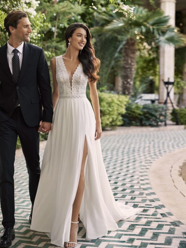 Couple souriant marchant main dans la main, la mariée portant une robe fluide avec un décolleté en V en dentelle et une fente élégante