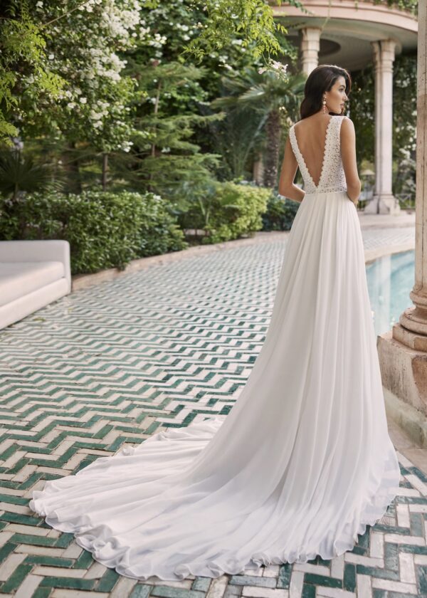 Mariée vue de dos portant une robe de mariée fluide avec un dos en V orné de dentelle, se tenant près d'une piscine dans un jardin luxuriant