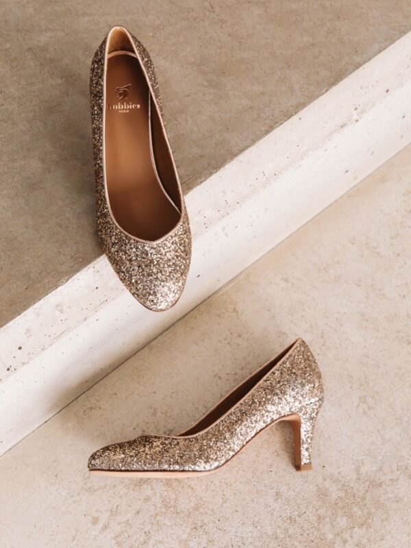 Vue de face d’une paire d’escarpins dorés pailletés posés sur un sol en béton texturé.