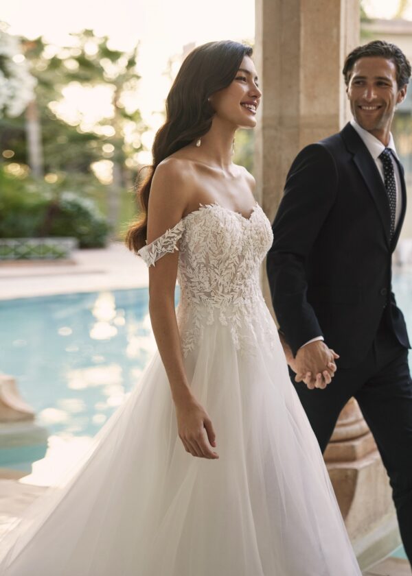 Mariée souriante en robe de mariée à épaules dénudées, main dans la main avec le marié, près d'une piscine