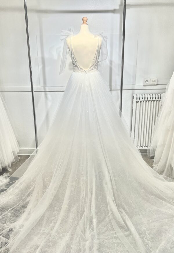 Robe de mariée princesse en tulle pailleté, avec un décolleté en V profond, un bustier orné de pierreries, un dos nu sublimé par des bretelles délicates et des nœuds en tulle aux épaules.