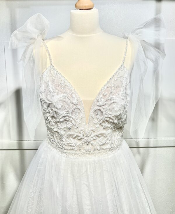 Robe de mariée princesse en tulle pailleté, avec un décolleté en V profond, un bustier orné de pierreries, un dos nu sublimé par des bretelles délicates et des nœuds en tulle aux épaules.