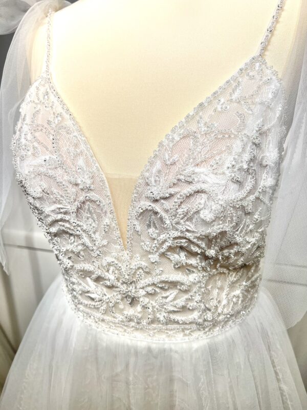 Robe de mariée princesse en tulle pailleté, avec un décolleté en V profond, un bustier orné de pierreries, un dos nu sublimé par des bretelles délicates et des nœuds en tulle aux épaules.