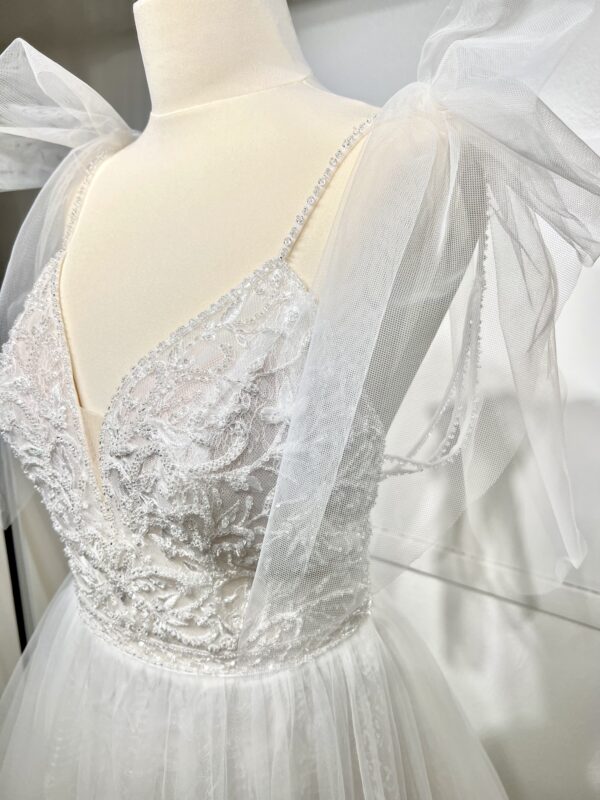 Robe de mariée princesse en tulle pailleté, avec un décolleté en V profond, un bustier orné de pierreries, un dos nu sublimé par des bretelles délicates et des nœuds en tulle aux épaules.