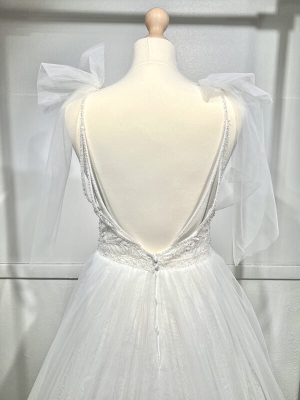 Robe de mariée princesse en tulle pailleté, avec un décolleté en V profond, un bustier orné de pierreries, un dos nu sublimé par des bretelles délicates et des nœuds en tulle aux épaules.
