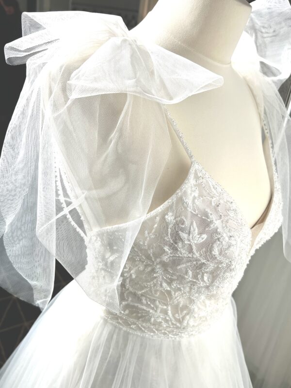 Robe de mariée princesse en tulle pailleté, avec un décolleté en V profond, un bustier orné de pierreries, un dos nu sublimé par des bretelles délicates et des nœuds en tulle aux épaules.