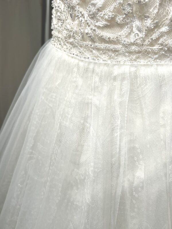 Robe de mariée princesse en tulle pailleté, avec un décolleté en V profond, un bustier orné de pierreries, un dos nu sublimé par des bretelles délicates et des nœuds en tulle aux épaules.