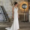 Une robe de mariée ajustée avec un décolleté illusion en dentelle, sans manches, et une traîne fluide. Le cadre industriel en arrière-plan met en valeur l'élégance contemporaine de la robe
