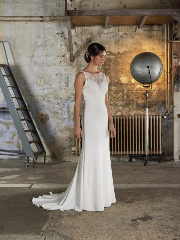 Une robe de mariée ajustée avec un décolleté illusion en dentelle, sans manches, et une traîne fluide. Le cadre industriel en arrière-plan met en valeur l'élégance contemporaine de la robe