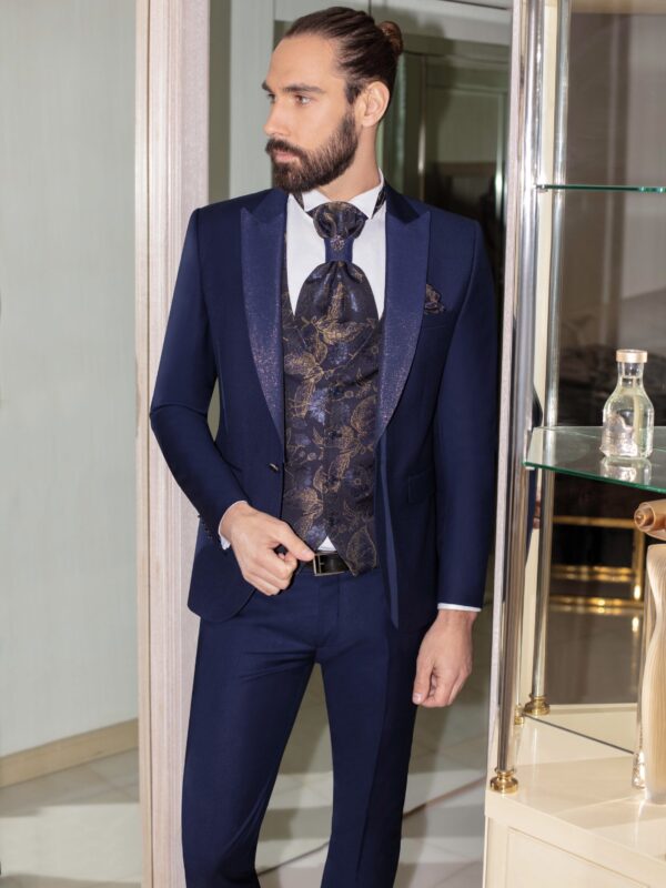 Homme vêtu d’un costume bleu marine profond avec des détails dorés scintillants, complété par un gilet et une lavallière richement ornés de motifs dorés, dans un intérieur élégant.