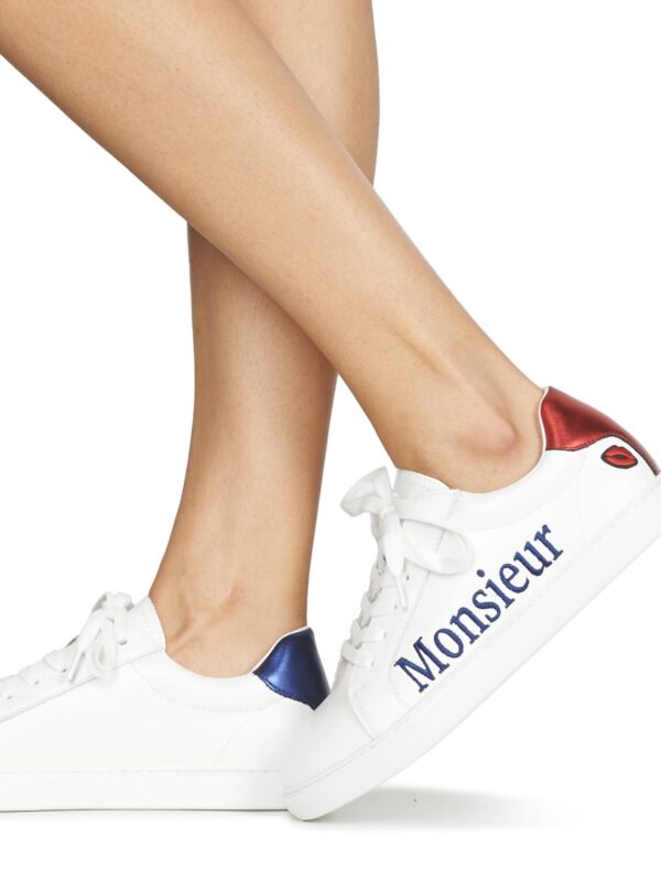 Une personne portant des baskets blanches avec les inscriptions « Monsieur » et des talons métallisés rouge et bleu.