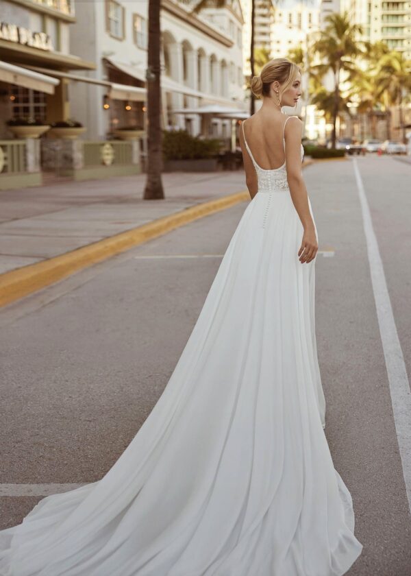 Vue arrière d'une mariée dans une robe fluide et épurée, ornée d'un dos décolleté et de détails subtils en dentelle, marchant dans une rue ensoleillée bordée de palmiers et de bâtiments modernes