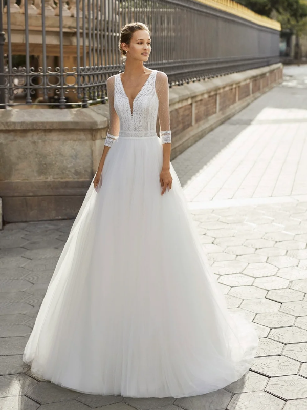Une femme élégante en robe de mariée blanche à manches longues en dentelle, un décolleté en V et une jupe en tulle volumineuse, posant devant une grille dorée.