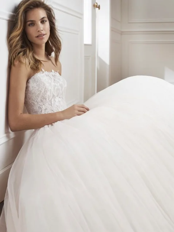 Mariée assise dans une robe de princesse volumineuse en tulle, avec un corsage en dentelle délicate, posant dans un intérieur élégant aux tons clairs et doux