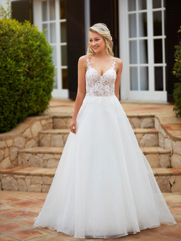 Une mariée souriante portant une robe blanche avec un corsage en dentelle florale, un décolleté en V et une jupe fluide en tulle, posant devant une villa élégante.