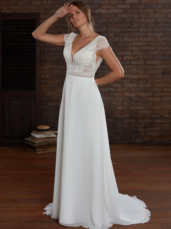 Une femme portant une robe de mariée fluide et élégante, caractérisée par un corsage en dentelle délicate, des manches transparentes et un décolleté plongeant, posant dans un cadre intérieur au style rustique