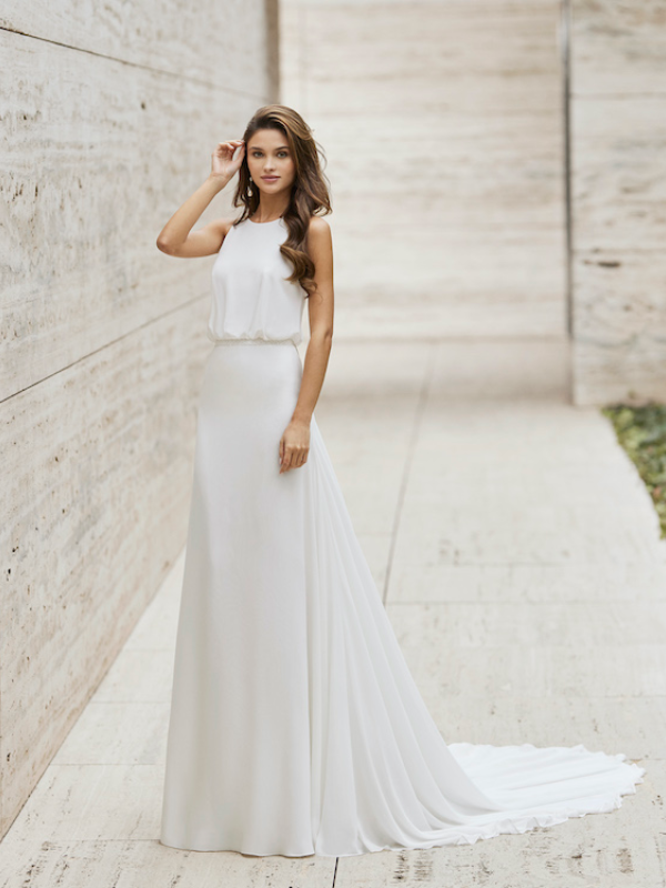 Une mariée portant une robe blanche minimaliste avec un haut fluide sans manches et une jupe droite, posant dans un couloir moderne en pierre.