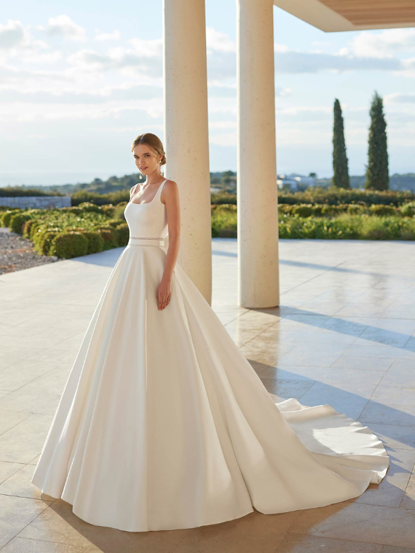Une mariée pose dans une élégante robe de mariée princesse à col carré et jupe volumineuse, devant un paysage moderne baigné de lumière.