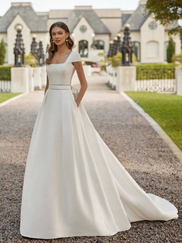 Une mariée élégante portant une robe de mariée princesse avec un col carré structuré et une jupe ample, posant dans une allée bordée d’un château majestueux.